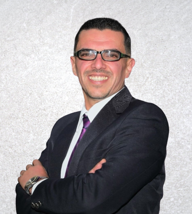 Man in a dark suit and purple tie smiling with arms crossed against a light gray textured background
